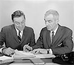 1960s TWO BUSINESSMEN SEATED AT DESK LOOKING AT FILE FOLDERS