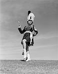 1950s WOMAN MAJORETTE IN UNIFORM WITH SHORT SKIRT BOOTS TALL FURRY HAT POSING OUTDOORS HOLDING A BATON