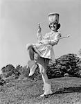 1960ER JAHRE LÄCHELND MAJORETTE IN UNIFORM MIT KURZEN ROCK STIEFEL HOHEN HUT UND ZWEI STÖCKCHEN POSIERT IM FREIEN