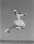 1960s WOMAN MAJORETTE WEARING BAND UNIFORM HOLDING BATON JUMPING INTO THE AIR