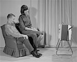 1960s COUPLE MAN WOMAN SITTING CHAIR WATCHING PORTABLE TELEVISION