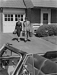 1950s COUPLE CARRYING LUGGAGE WALKING TOWARDS CONVERTIBLE CAR IN DRIVEWAY