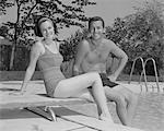 1960s MAN WOMAN COUPLE SITTING ON DIVING BOARD ON SIDE OF SWIMMING POOL