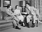 1950s COUPLE SITTING ON COUCH TALKING TO SMILING INSURANCE SALESMAN HANDING THEM A POLICY