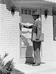 1950s SALESMAN WEARING SUIT TIE HAT CARRYING BRIEFCASE RINGING SUBURBAN HOUSE DOORBELL