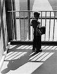 1950s SCHOOLBOY WITH BOOKS UNDER ARM LOOKING OUT WINDOW BETWEEN BARS OF RAILING SHADOW CAST BEHIND HIM