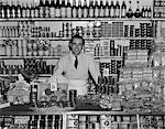 HOMME SOURIANT DES ANNÉES 1940 ÉPICIER DEBOUT DERRIÈRE LE COMPTOIR REMPLI DE DIVERS PRODUITS ALIMENTAIRES SUR LES BOÎTES ET PAQUETS INTÉRIEUR ÉPICERIE