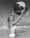 1950s SIDE VIEW OF BLONDE IN WHITE BIKINI STANDING IN POOL IN WAIST-HIGH WATER HOLDING BEACH BALL IN AIR
