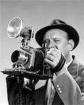 1950s PORTRAIT OF PHOTOGRAPHER MAN IN SUIT AND FEDORA HAT SQUINTING EYE TO LOOK THROUGH VIEWFINDER OF PRESS CAMERA