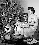 1940s FAMILY FATHER SON DAUGHTER SITTING TOGETHER IN FRONT OF CHRISTMAS TREE MOTHER OPENING PRESENT