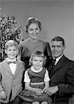 1950s - 1960s FAMILY PORTRAIT SMILING FATHER MOTHER DAUGHTER SON SITTING TOGETHER IN FRONT OF INDOOR CHRISTMAS TREE