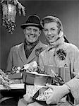 ANNÉES 1950 COUPLE MARI ET FEMME TRANSPORTANT BRASSÉE DE COLIS DE NOËL SOURIANT DEBOUT DANS LA NEIGE SOUS LE PORCHE DE LUMIÈRE EN PLEIN AIR