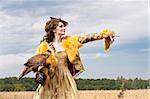 The woman in a beautiful old style dress with falcon has a rest before hunting in yellow field.