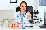 Smiling female doctor sitting at office table and showing victory gesture