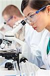 A Chinese Asian female medical or scientific researcher or doctor using a microscope in a laboratory with her colleague out of focus behind her.