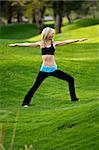 Beautiful blond young woman practicing yoga in the park on a green lawn with trees in the distance. She is in Warrior One pose.