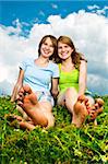 Two young teenage girl friends sitting barefoot on summer meadow