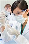 A Chinese Asian female medical or scientific researcher or doctor using looking at a test tube of clear liquid in a laboratory with her colleague out of focus behind her.