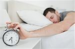 Sleeping young man being awakened by an alarm clock in his bedroom