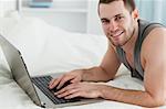 Handsome man using a laptop while lying on his belly in his bedroom