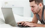 Serious man using a laptop while lying on his belly in his bedroom