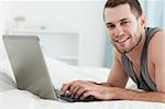 Happy man using a laptop while lying on his belly in his bedroom