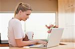 Woman using a laptop while drinking a cup of a tea in her kitchen