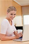 Portrait of a woman using a laptop while drinking a cup of a coffee in her kitchen