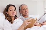 Smiling woman reading a book while her husband is reading the news in their bedroom