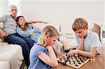 Siblings playing chess in front of their parents in a living room