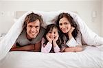 Smiling parents lying under a duvet with their daughter in their bedroom