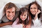 Happy parents lying under a duvet with their daughter in their bedroom