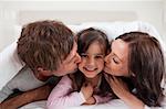 Parents kissing their daughter while lying under a duvet