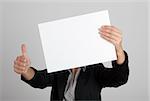 Woman showing a blank paper sheet in front of her head and making a thumbs up gesture with the right hand