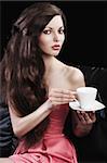 sophisticated young pretty brunette in pink elegant dress sitting on a black sofa and tasting a cup of tea. she looks in to the lens with  three quarters turned faces  and takes a cup of tea with both hands