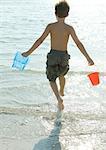 Boy running into surf with buckets, rear view