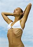 Woman in bikini stretching with hands behind head and eyes closed, low angle view