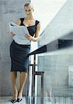 Businesswoman leaning against glass guard rail, reading newspaper, full length