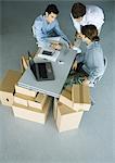 Drei männliche Kollegen gruppieren sich um Handy auf Schreibtisch, umgeben von Kartons