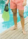 Man standing by edge of swimming pool, holding flippers, waist down