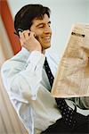 Businessman using phone and holding up financial section of newspaper, smiling