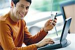 Man using laptop and dialing cell phone, smiling at camera