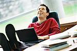 Man sitting at desk with feet up, holding laptop on lap, laughing