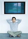 Man sitting on floor under wide screen TV, hands behind head and eyes closed