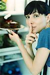 Woman taking piece of cake from refrigerator, looking at camera with finger over lips
