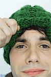Young man lifting up edge of knit hat, looking at camera, close-up