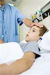 Boy lying in hospital bed, doctor taking temperature and feeling forehead