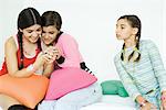 Three young female friends sitting on sofa, two looking at cell phone together while third watches