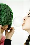 Two young female friends, one blowing bubble with chewing gum while the other pulls hat over eyes