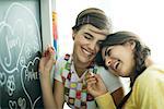 Young female friends writing names in hearts on chalkboard, laughing